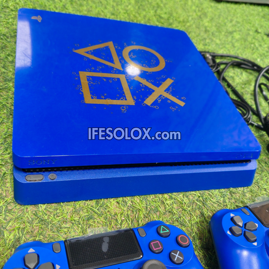 A blue Sony PlayStation 4 console with gold PlayStation logo graphics on the lid, shown on a green grass-like surface with two matching blue controllers partially visible.