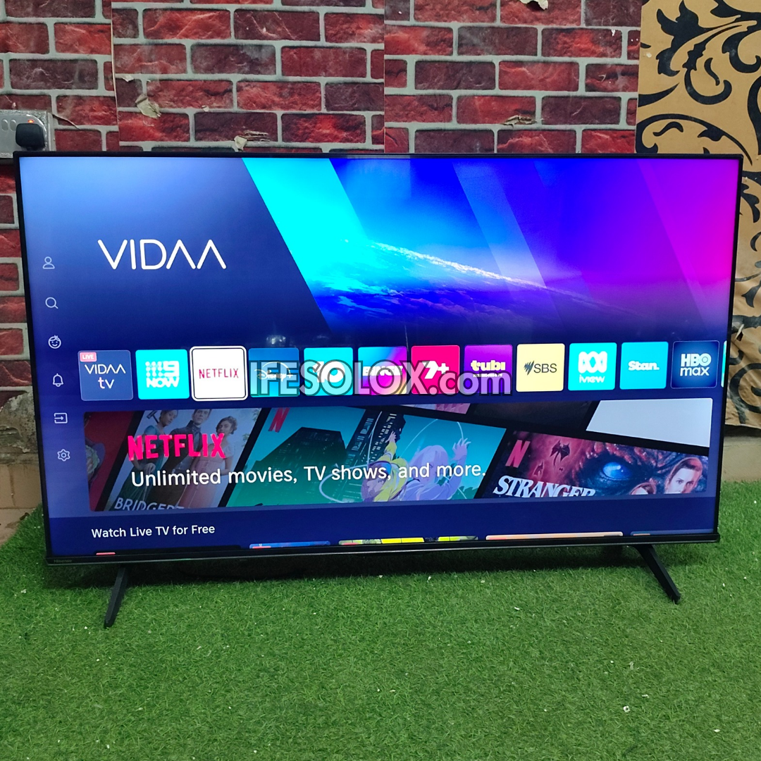 A large Hisense VIDAA smart TV with a frameless bezel and stand, displaying a colorful Netflix and streaming app interface, set against a brick wall and artificial grass background.