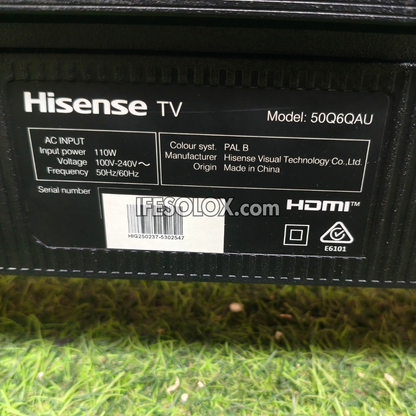 Close-up of the back of a Hisense TV power supply or receiver, showing printed specifications, model 50Q6QAU, HDMI text, and a barcode label on a black surface above artificial green grass.
