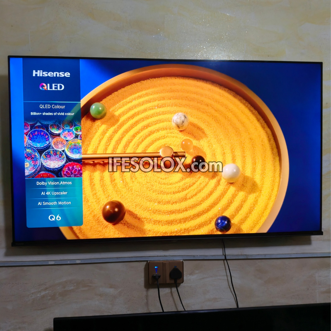 A Hisense QLED TV Q6 mounted on a beige wall, displaying a colorful screen with a zen garden image and a blue sidebar. The wall behind the TV is slightly textured, and an outlet with plugged-in devices is visible below the TV.