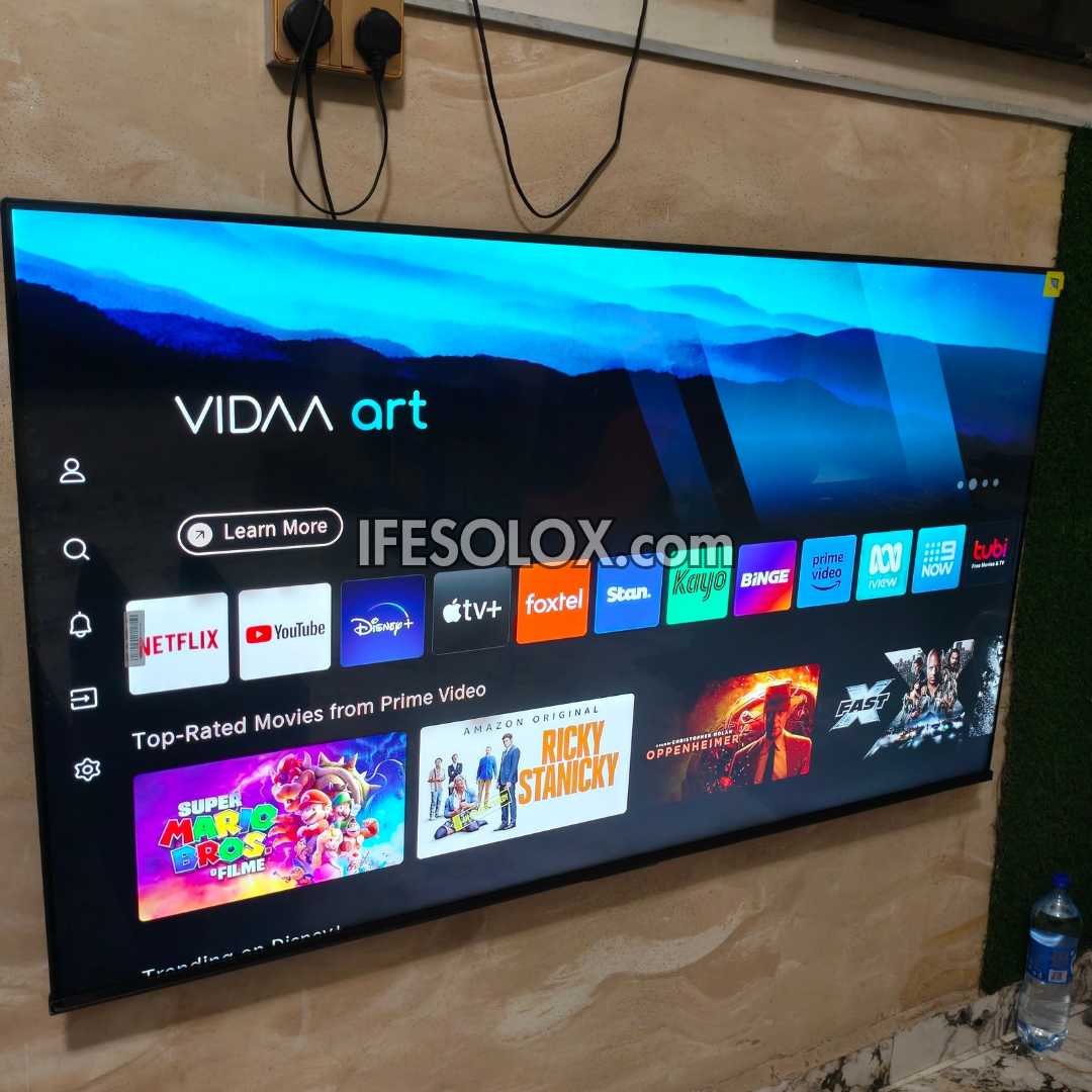 A large Hisense VIDAA art smart TV mounted on a beige stone wall, displaying a colorful app grid with Netflix, YouTube, Disney+, Apple TV+, Foxtel, Stan, Bing, HBO, and Prime Video. A watermark 'IFESOLOX.