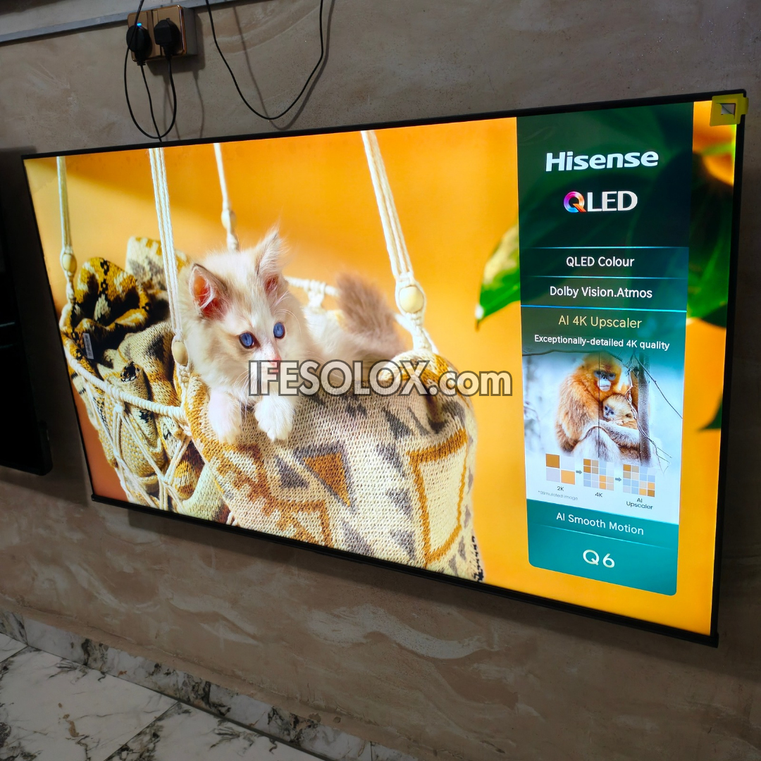 A Hisense QLED TV mounted on a beige marble-patterned wall, displaying a colorful screen with a fluffy white kitten in a woven basket and a vertical feature panel reading Hisense QLED, QLED Colour, Dolby Vision Atmos, AI 4K Upscaling, and Q6.