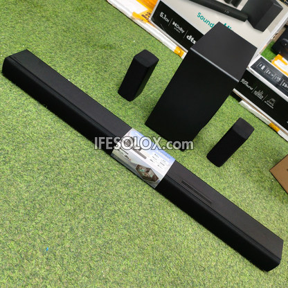 A black 5.1 soundbar speaker system with a subwoofer and two mid-range speakers, displayed on a green textured carpet with product boxes in the background.