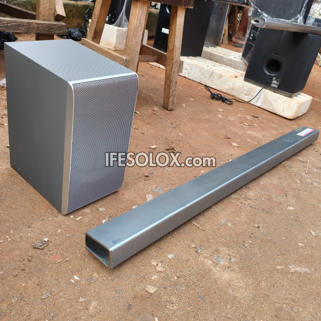 A silver LG metal soundbar and matching subwoofer displayed outdoors on a dirt ground, with a wooden table and other equipment in the background. A watermark 'IFESOLOX.