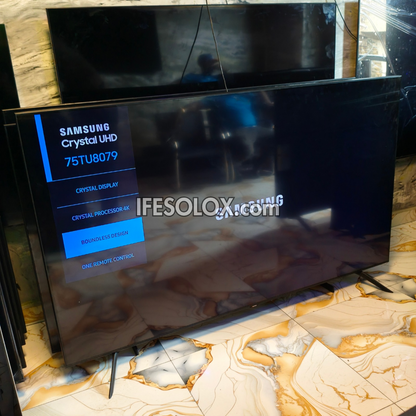 A Samsung Crystal UHD 75TU8079 television shown at an angle in a showroom, with its screen turned on displaying the Samsung Crystal Display interface. The TV is resting on a polished beige and gold marble-patterned floor, with other TVs and a mirror visible in the background.