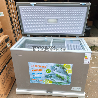 A Snowsea brand 250L top-freezer chest freezer with a silver-grey metal front, glass top, and open white interior lid, photographed outdoors on a concrete floor next to stacked cardboard boxes.
