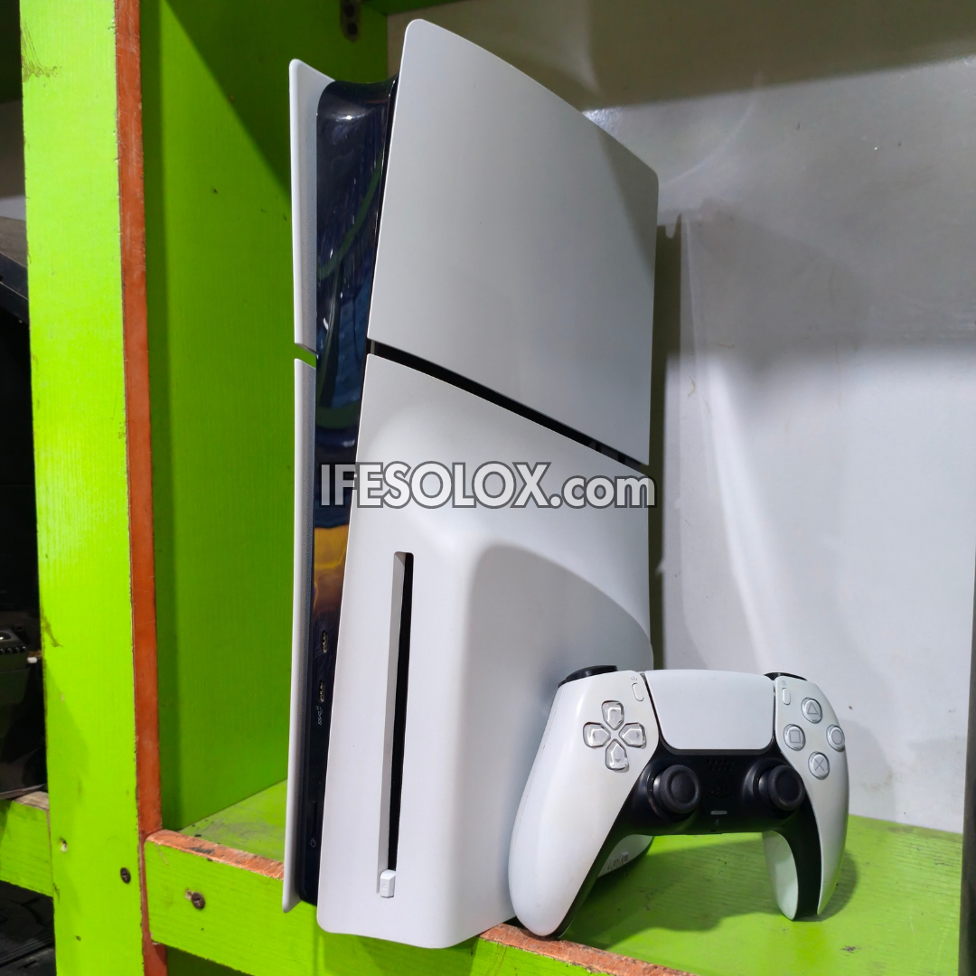 White PlayStation 5 Slim console standing vertically next to a matching white and black DualSense controller on a green shelf. A semi-transparent watermark reading 'IFESOLOX.