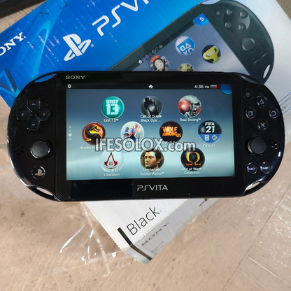 A Sony PlayStation Vita handheld console in black, shown on top of its blue retail box and a white paper insert on a light wood surface. The screen is on and displaying the PS Vita home screen with various app icons.