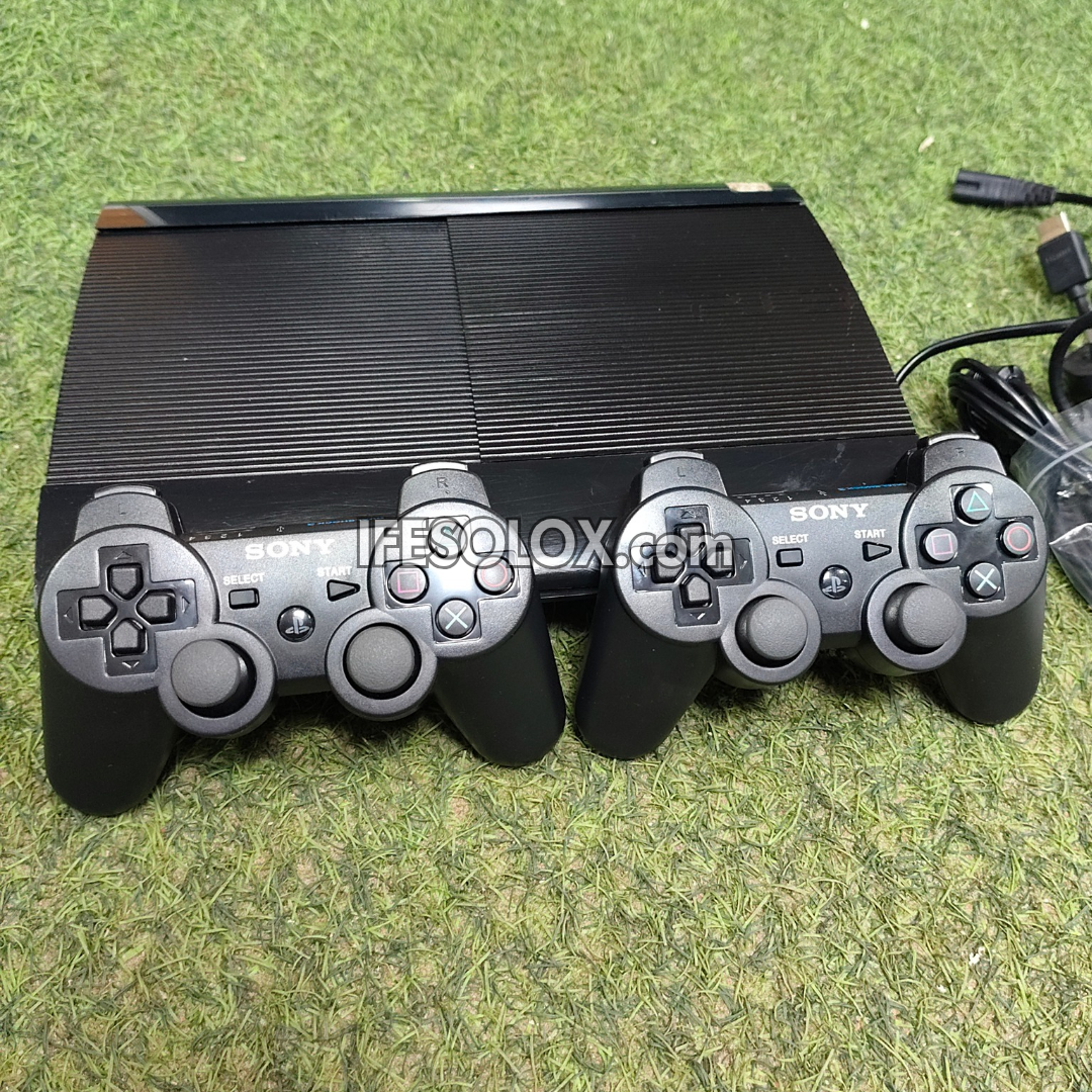 A Sony PlayStation 3 console with two black DualShock 3 wireless controllers placed in front of it, photographed on a green grass-like surface. A USB cable is visible at the upper right, and a large 'IFESOLOX.