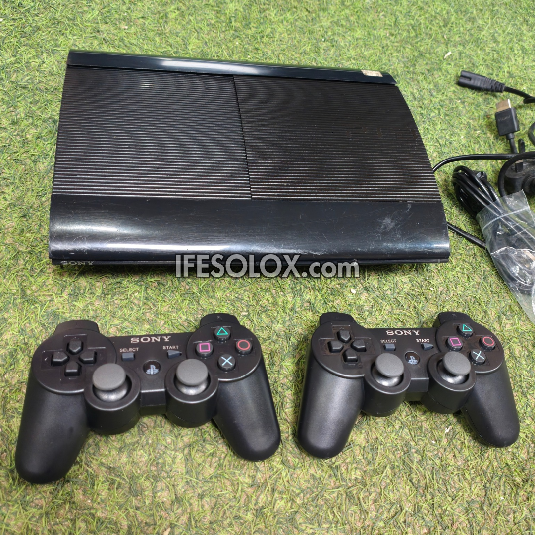 A black Sony PlayStation 3 console shown with two black DualShock wireless controllers on a green grass-like surface, with cables visible on the right and a visible IFESOLOX.com watermark across the center.