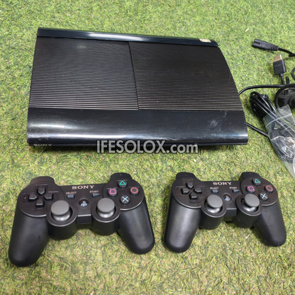 A black Sony PlayStation 3 console shown with two black DualShock wireless controllers on a green grass-like surface, with cables visible on the right and a visible IFESOLOX.com watermark across the center.