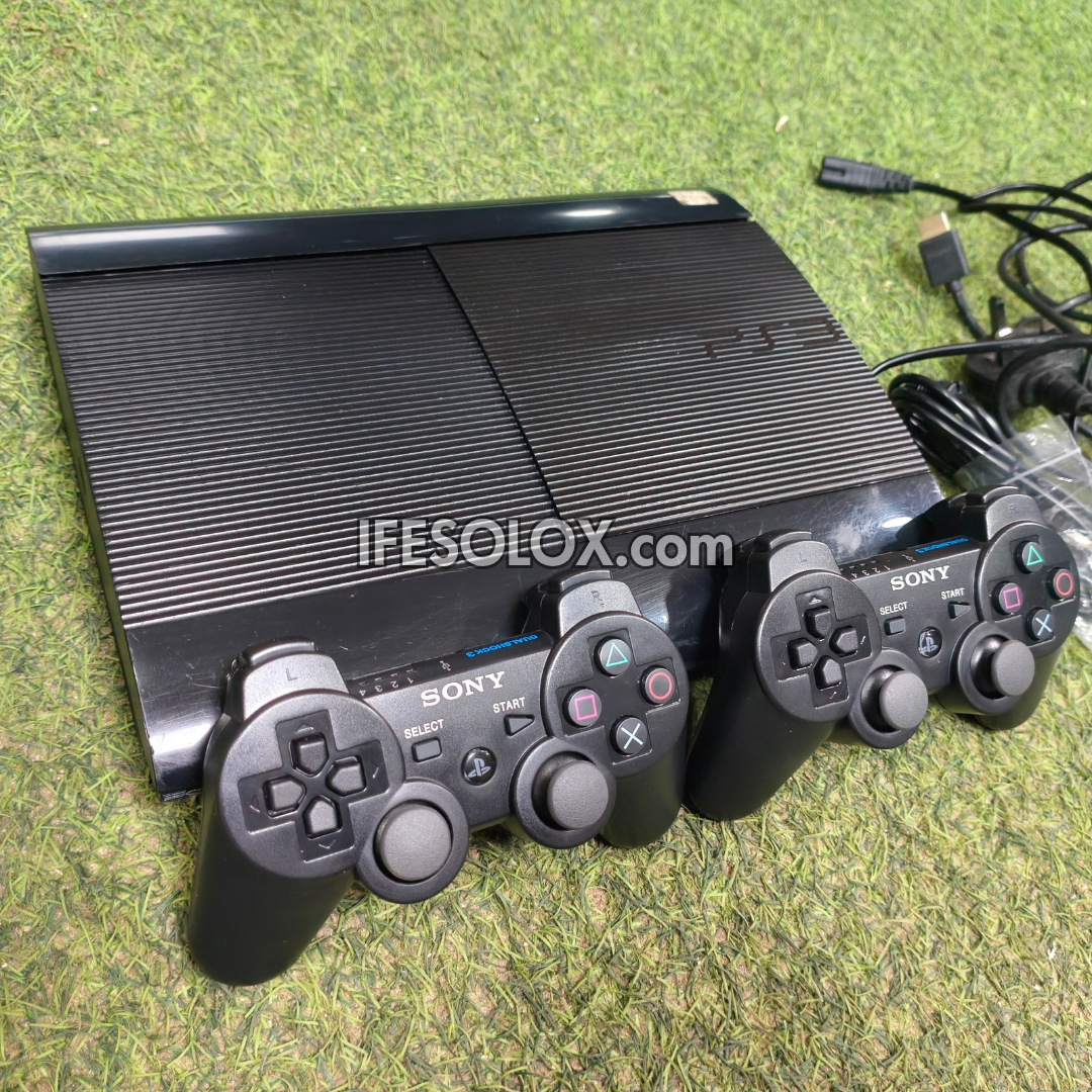 A black Sony PlayStation 3 console with two matching controllers placed in front of it on a green grass-like surface. Coiled cables and accessories are visible on the right side, and a semi-transparent 'IFESOLOX.