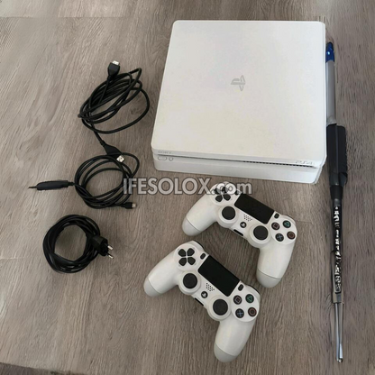 Sony Playstation 4 (PS4) Slim 1TB White Game Console with two DUALSHOCK 4 Controllers, USB charging cables, HDMI cable, and power cable laid out on a floor.