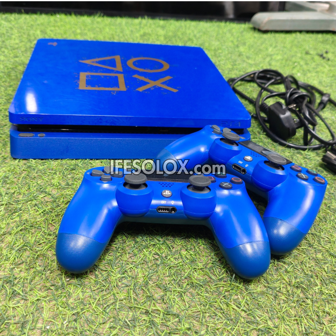A PlayStation 4 console in royal blue with two matching blue controllers and a power cable, placed on artificial green grass. The PlayStation logo is visible on the console lid, and a watermark reading 'IFESOLOX.