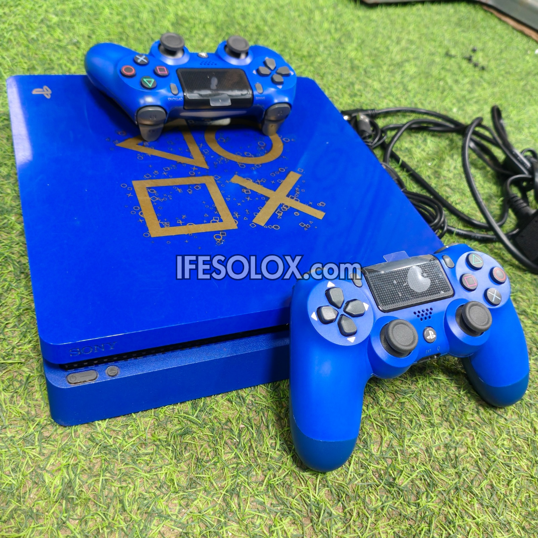A limited edition custom blue PlayStation 4 Slim console with gold PlayStation logo graphics, accompanied by two blue DualShock 4 controllers, placed on green artificial grass.