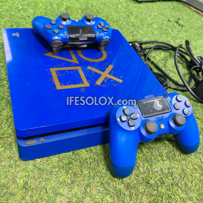 A limited edition custom blue PlayStation 4 Slim console with gold PlayStation logo graphics, accompanied by two blue DualShock 4 controllers, placed on green artificial grass.