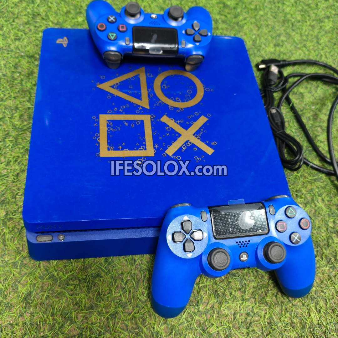 A blue PlayStation 4 Slim console with a matching blue controller on top, another blue controller in front, and a black power cable to the right, photographed on artificial green grass. The console lid has gold PlayStation logo symbols, and a watermark reading IFESOLOX.
