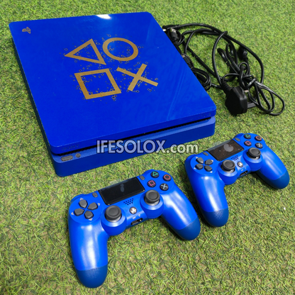 A PlayStation 4 console in a glossy royal blue finish with gold PlayStation logo symbols, shown with two matching blue wireless controllers and a power cable on a green grass-like surface. A visible watermark reads IFESOLOX.