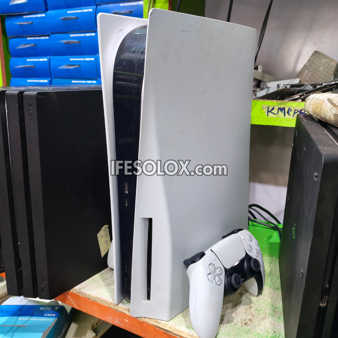 A white PlayStation 5 console with a matching white and black DualSense controller, shown alongside other PS4 pro consoles on a cluttered shelf. A large 'IFESOLOX.